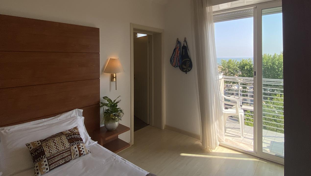Camera luminosa con letto, balcone vista mare e racchette appese al muro.